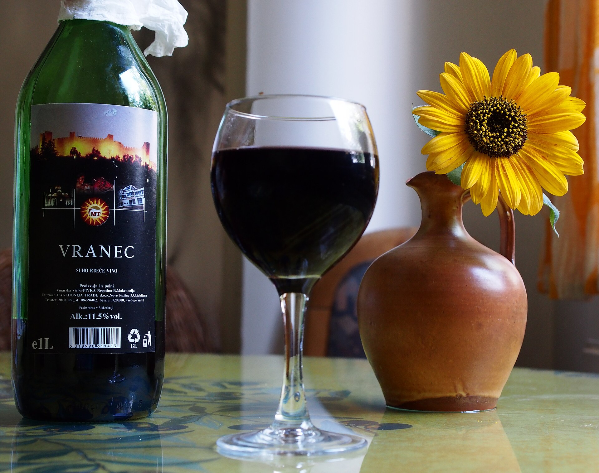 En flaska makedonskt Vranec-vin bredvid ett glas med det mycket mörka röda vinet på ett bord med en solros i en vas.
