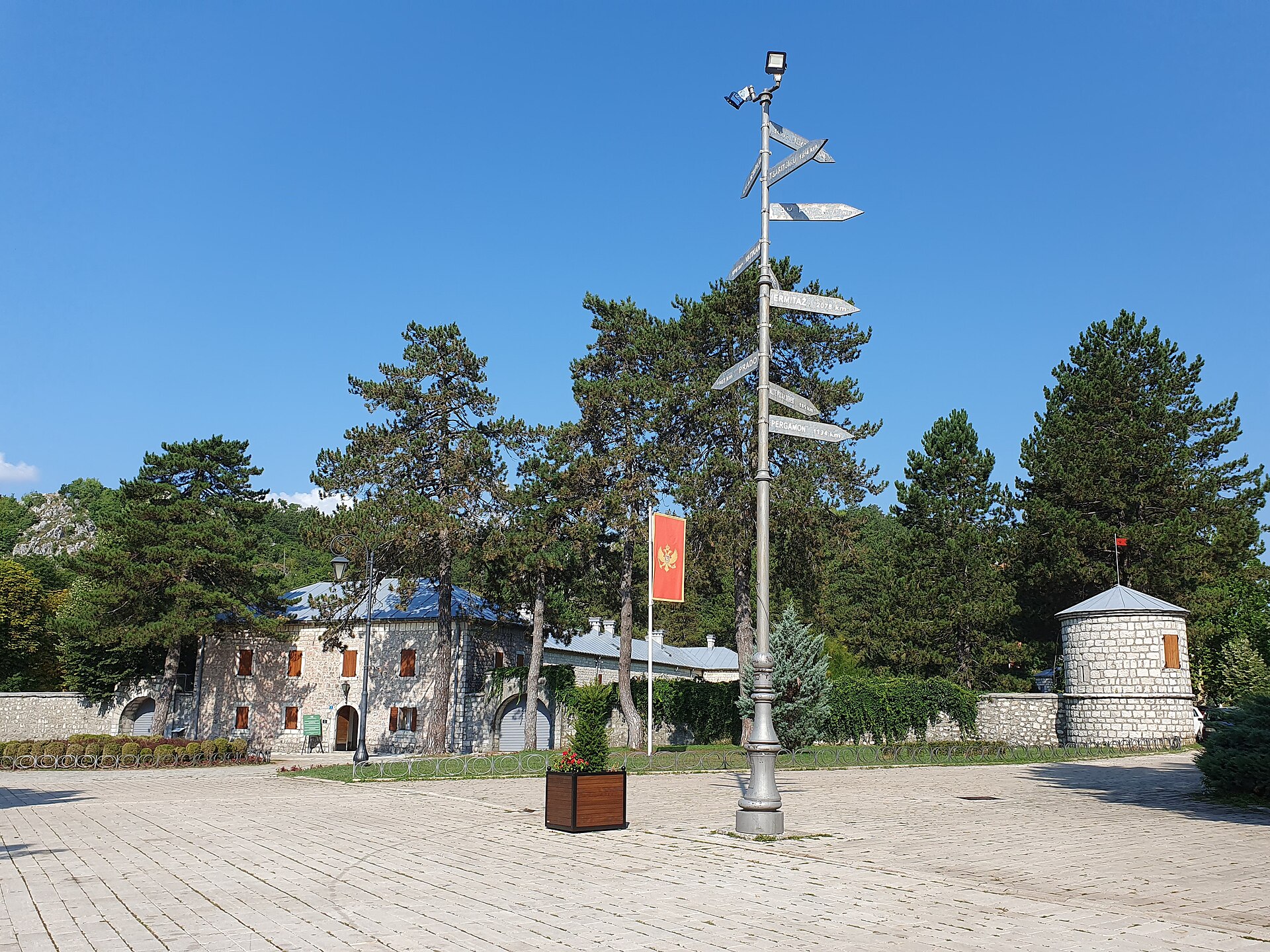En öppen stensatt plats framför en historisk stenbyggnad med en hög vägvisare i metall under en klarblå himmel.