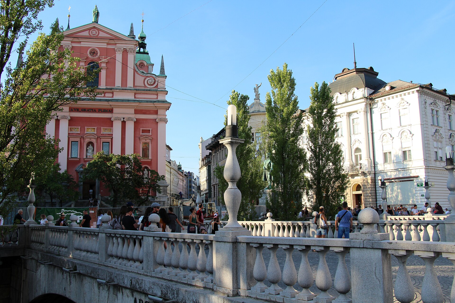 Fotgängare promenerar över Trippelbron i centrala Ljubljana med den rosa Franciskankyrkan i bakgrunden under en solig dag.