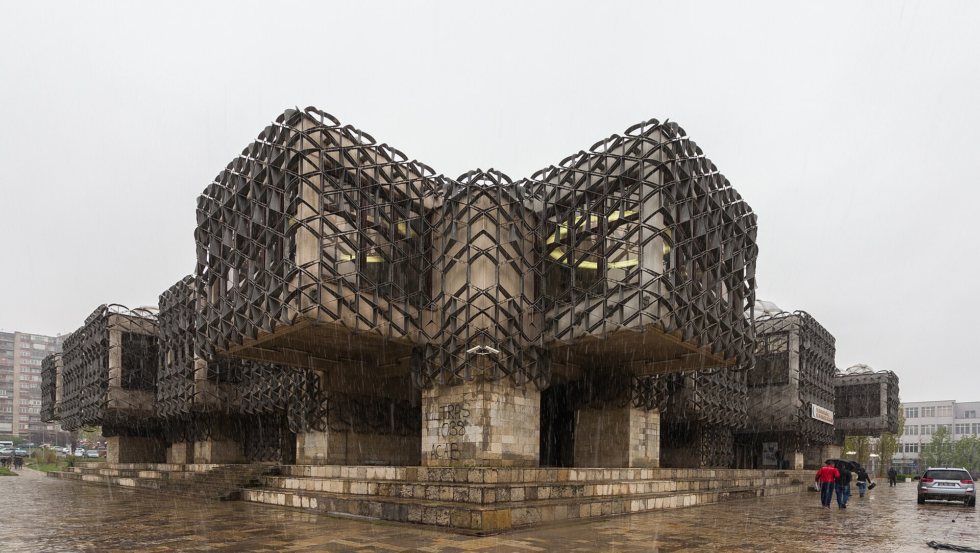 Kosovos nationalbibliotek i Pristina med sin karakteristiska fasad av metallnät, fotograferat en regnig dag med förbipasserande.