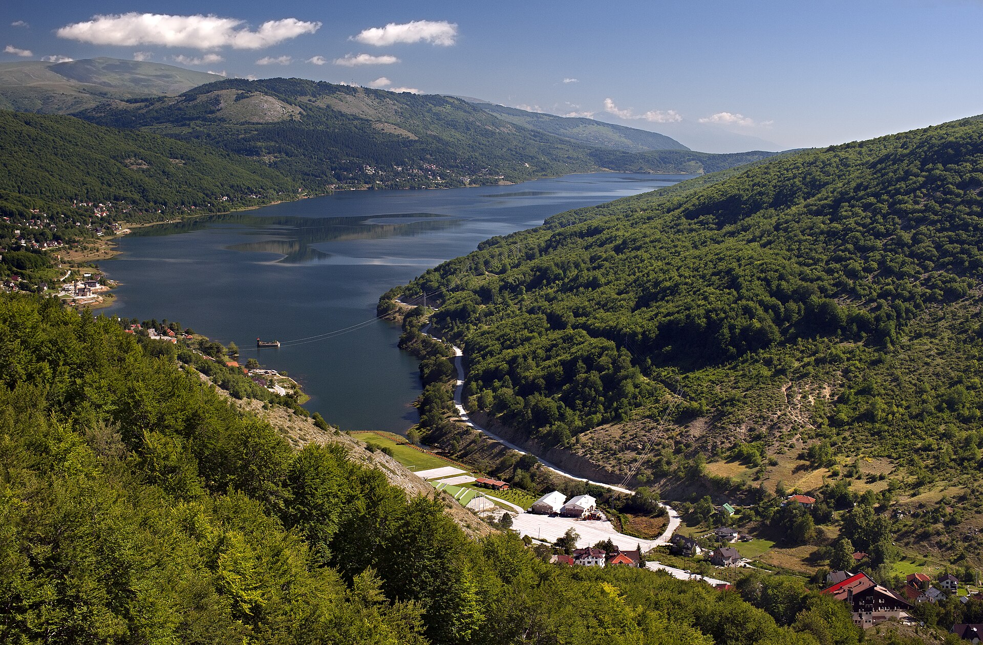 En vacker panoramavy över den blå Mavrovosjön omgiven av lummiga gröna skogsklädda bergssluttningar och små hus i bergsbyn.