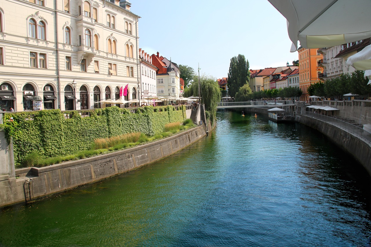En solig vy över floden Ljubljanica i Ljubljana, kantad av historiska byggnader och uteserveringar under vita parasoll.
