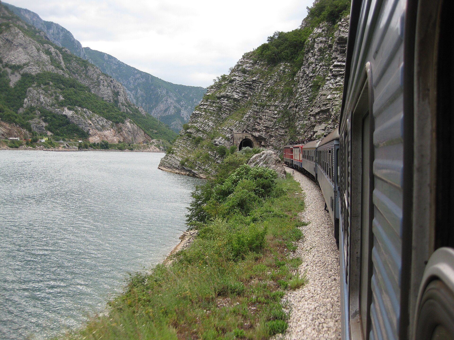 Ett tåg slingrar sig fram längs en turkos flod och branta bergssidor i en dalgång på Balkan.