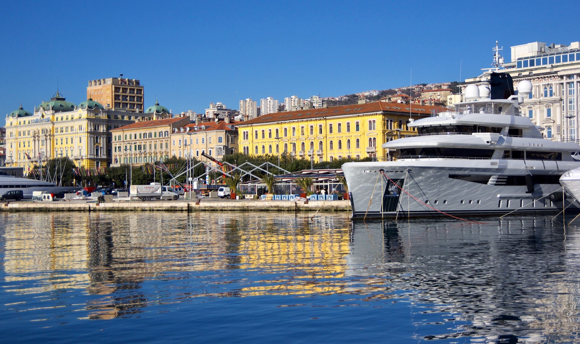 Färgglada byggnader i Rijeka speglar sig i det blå hamnvattnet bredvid en stor vit yacht vid kajen.