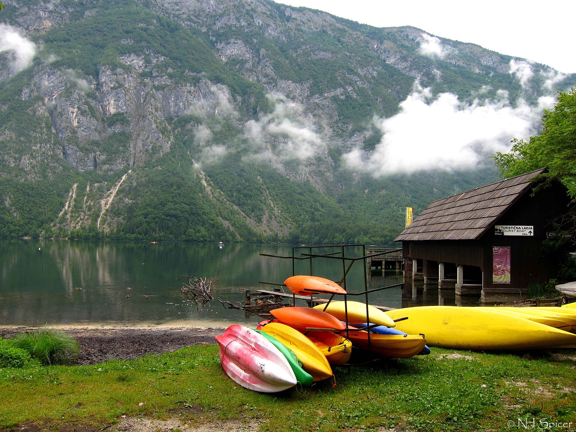 Bohinjsjön i Triglavs nationalpark med brant alpvägg, moln längs bergssidan och kajaker uppdragna på stranden.