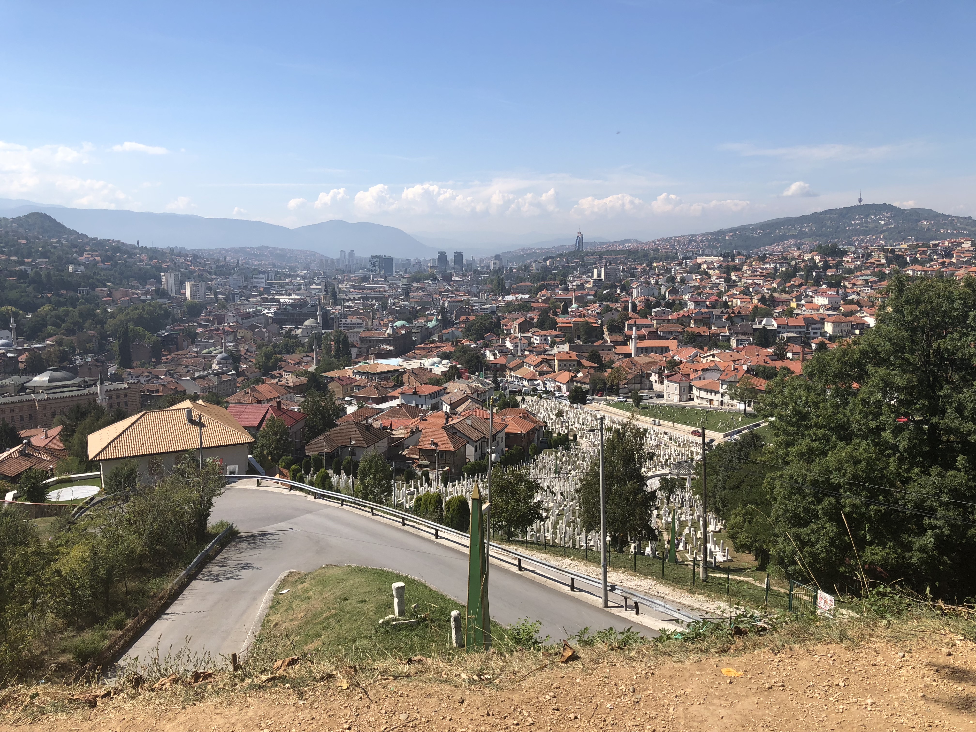 Panoramavy över Sarajevo från en höjd med vita gravstenar i förgrunden och stadens bostadskvarter och berg i bakgrunden.