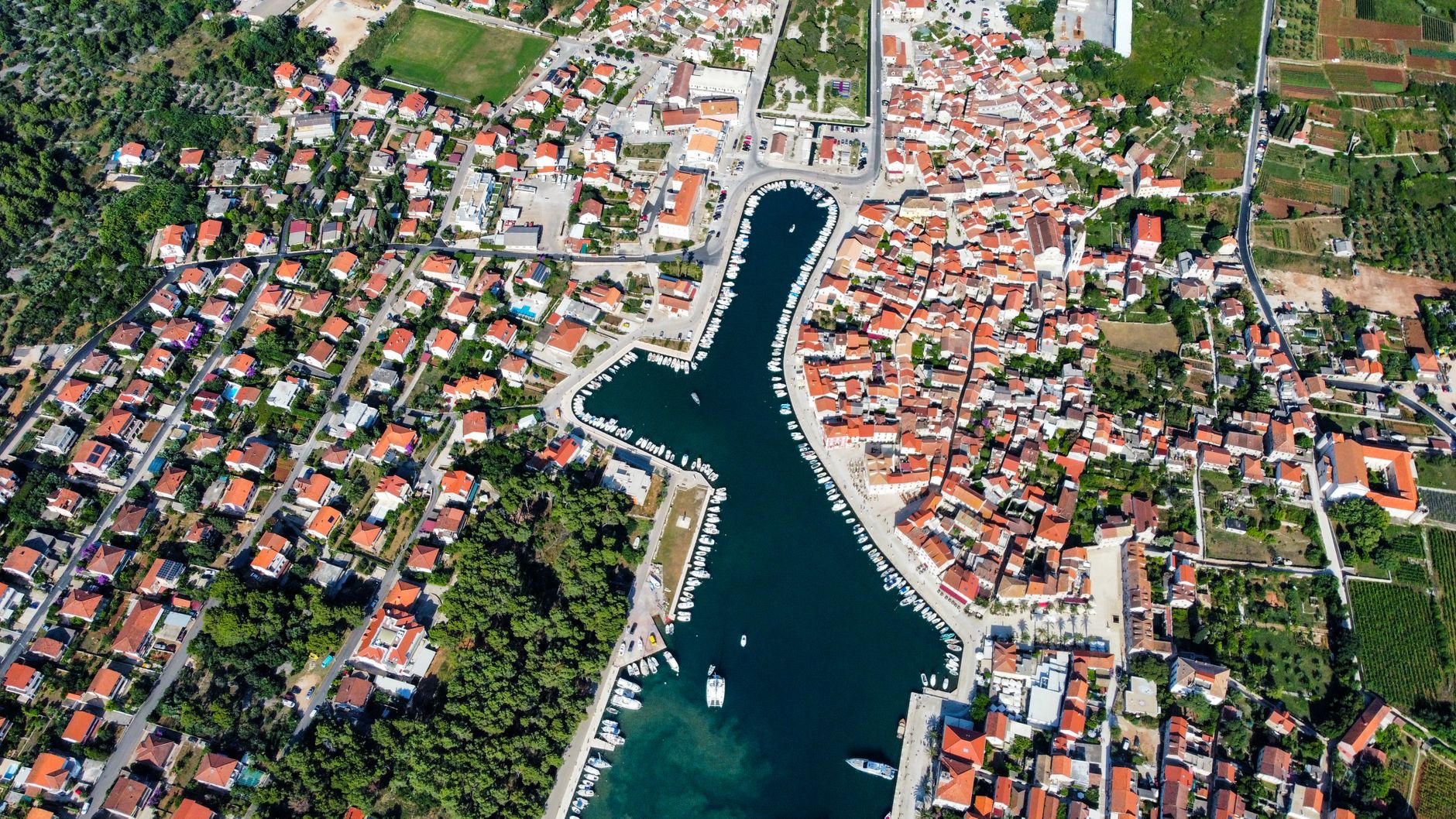 Ett vackert flygfoto över den långsmala hamnen i Stari Grad på ön Hvar, omgiven av hus med röda tak och lummig grönska.