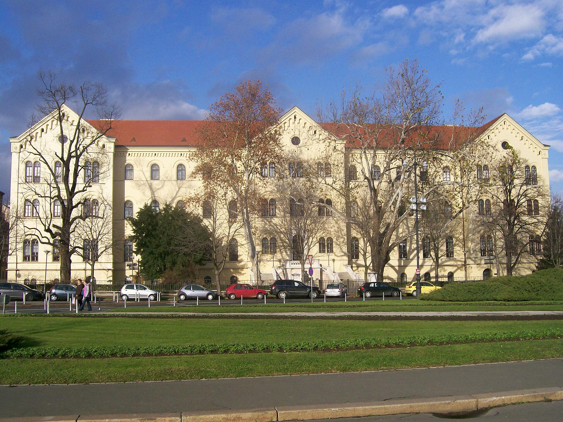 Universitetet i Zagrebs gula huvudbyggnad omgiven av träd under en blå himmel med vita moln.