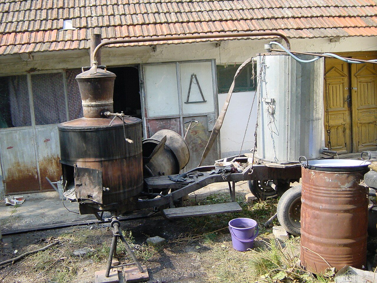 Traditionell kopparpanna för rakijabränning på en gårdsplan, med plommon och tunnor bredvid.