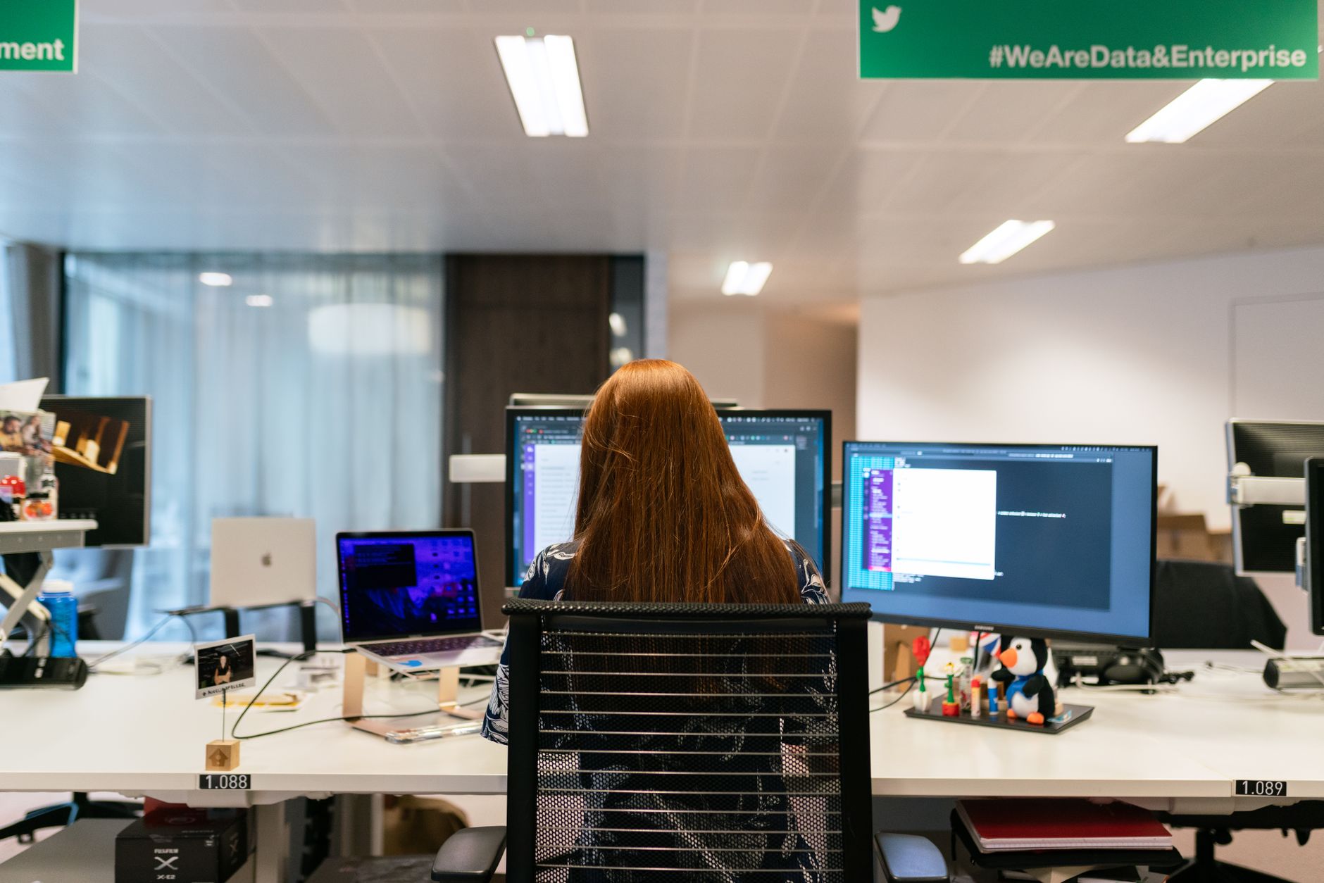 En datatekniker sedd bakifrån framför flera bildskärmar på ett ljust kontor.