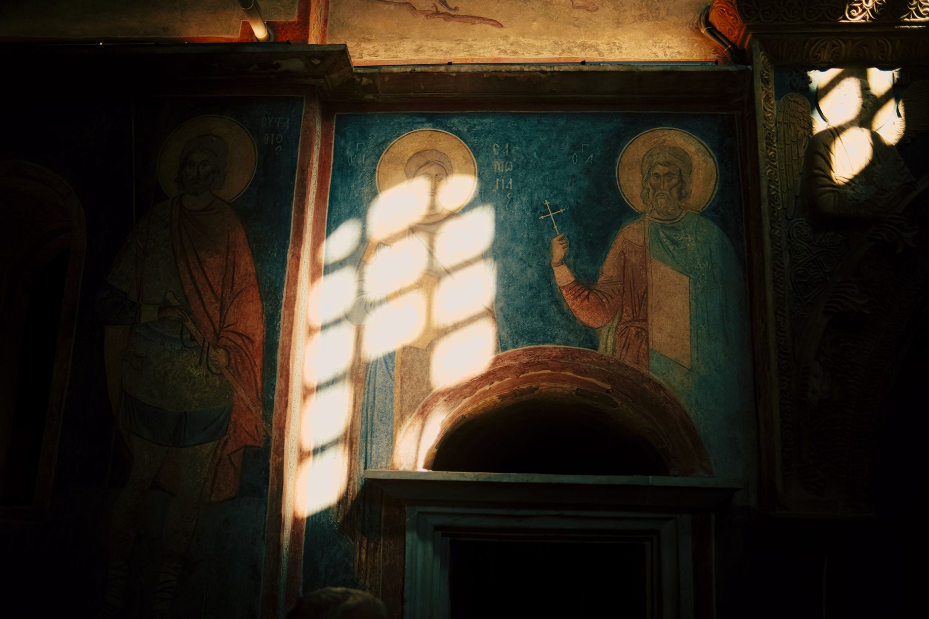 Medeltida fresker på en kyrkovägg i Visoki Dečani, där solljus faller över målade helgon och en gestalt som håller i ett svärd.
